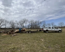 A white truck is parked with a trailer connected to it. Large pieces of lumber are being loaded on the back.