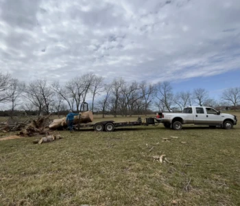 A white truck is parked with a trailer connected to it. Large pieces of lumber are being loaded on the back.